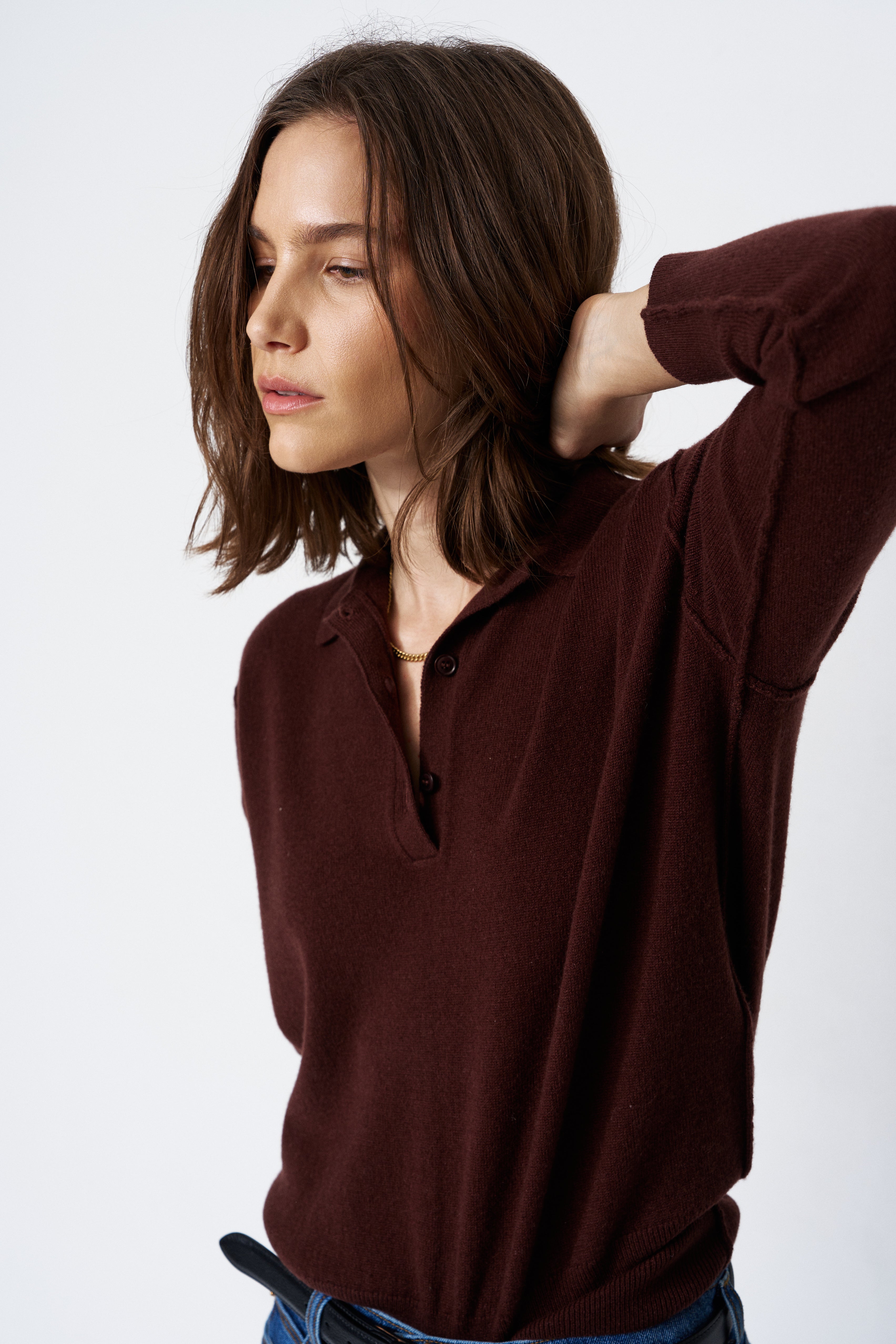 A woman wearing a dark brown long-sleeve knit polo shirt with a partial button placket, paired with blue jeans, photographed against a clean white studio background.