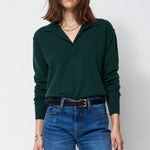 A woman modeling a dark green long-sleeve polo-neck sweater and blue wide-leg jeans with a black belt, standing against a plain white studio background.