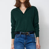 A woman modeling a dark green long-sleeve polo-neck sweater and blue wide-leg jeans with a black belt, standing against a plain white studio background.