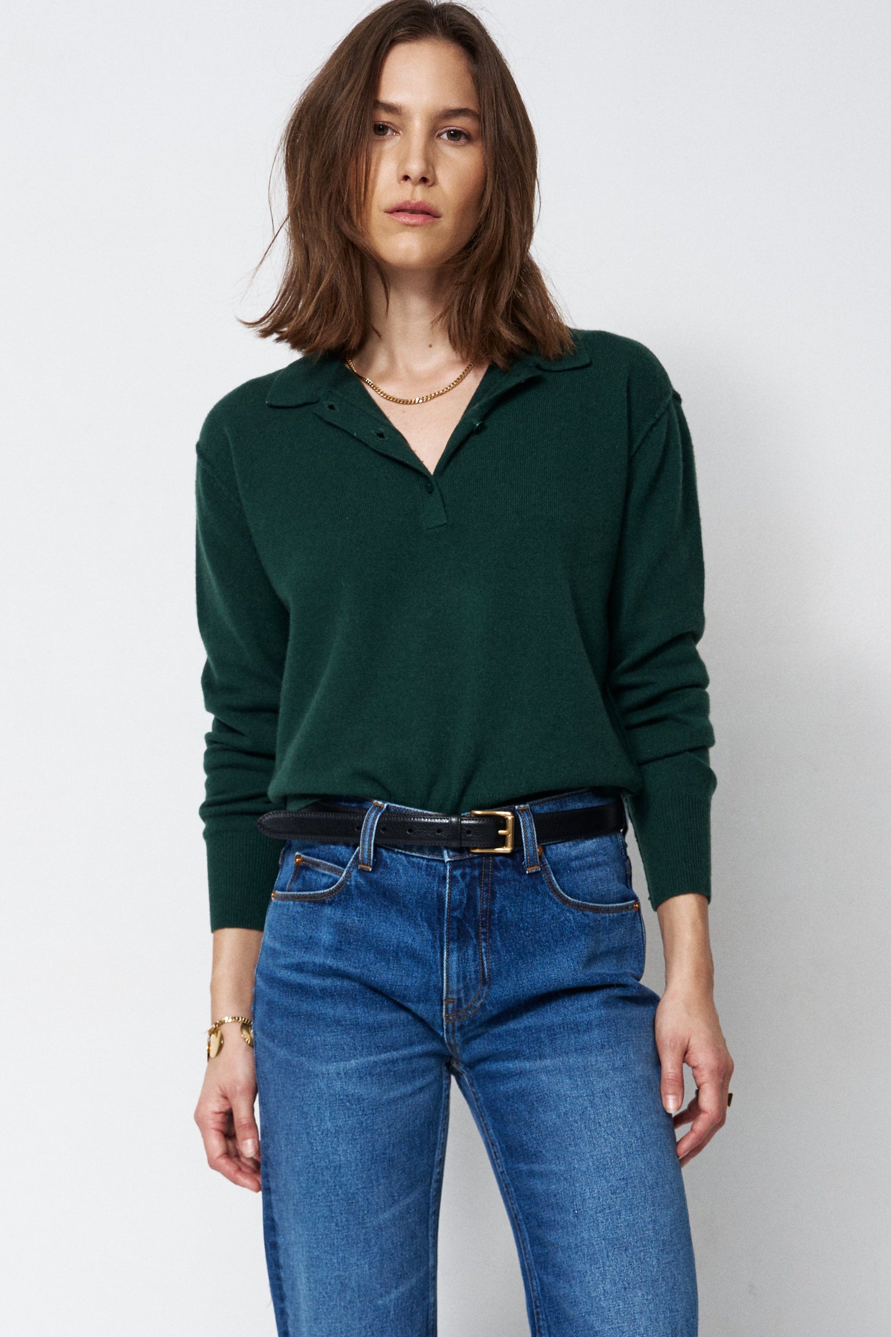 A woman modeling a dark green long-sleeve polo-neck sweater and blue wide-leg jeans with a black belt, standing against a plain white studio background.