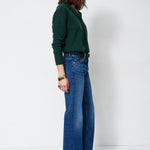 A woman stands in profile wearing a dark green long-sleeve top and blue wide-leg jeans against a plain white studio background.