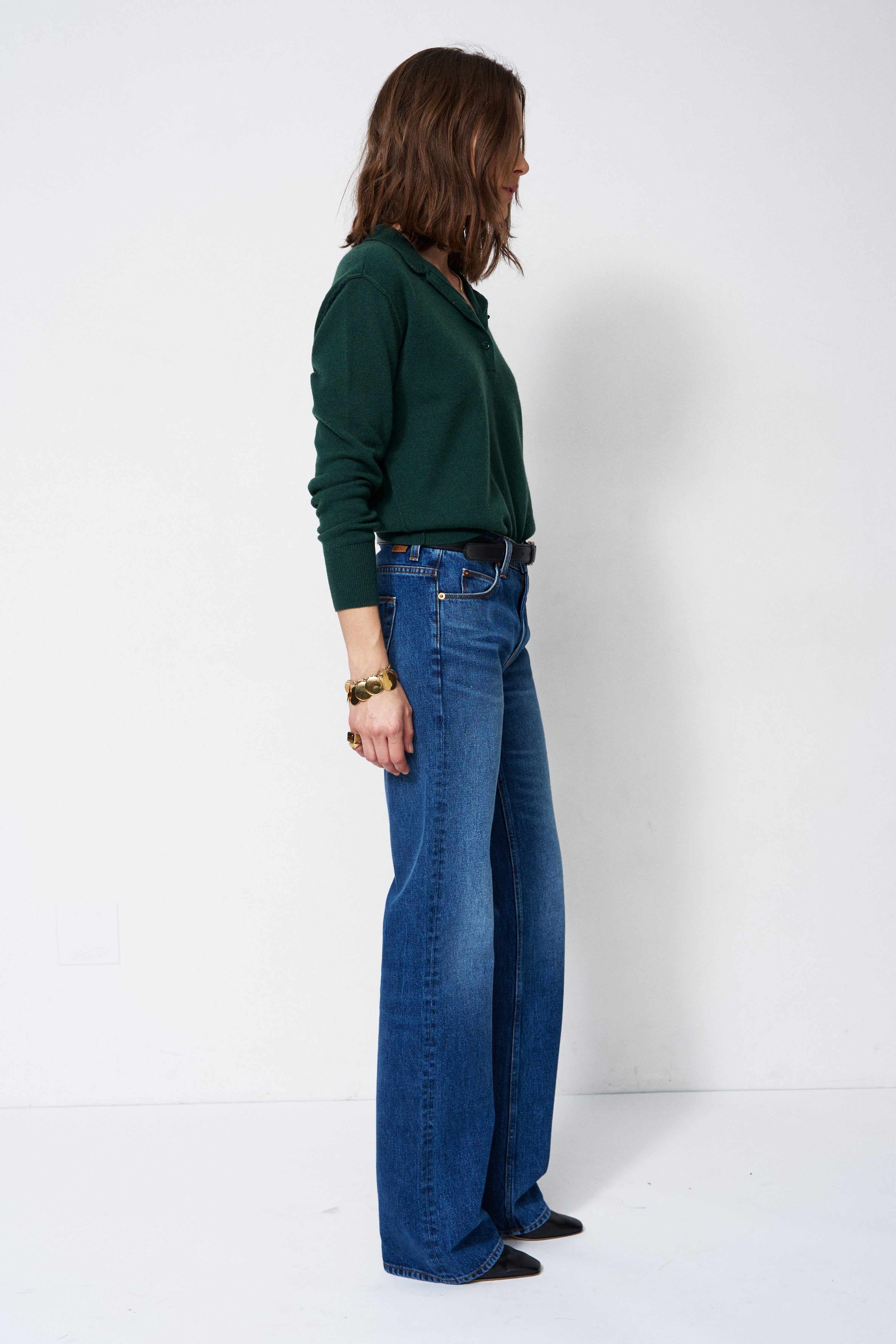A woman stands in profile wearing a dark green long-sleeve top and blue wide-leg jeans against a plain white studio background.