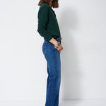 A woman stands in profile wearing a dark green long-sleeve top and blue flare jeans against a plain white studio background.