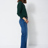 A woman stands in profile wearing a dark green long-sleeve top and blue flare jeans against a plain white studio background.