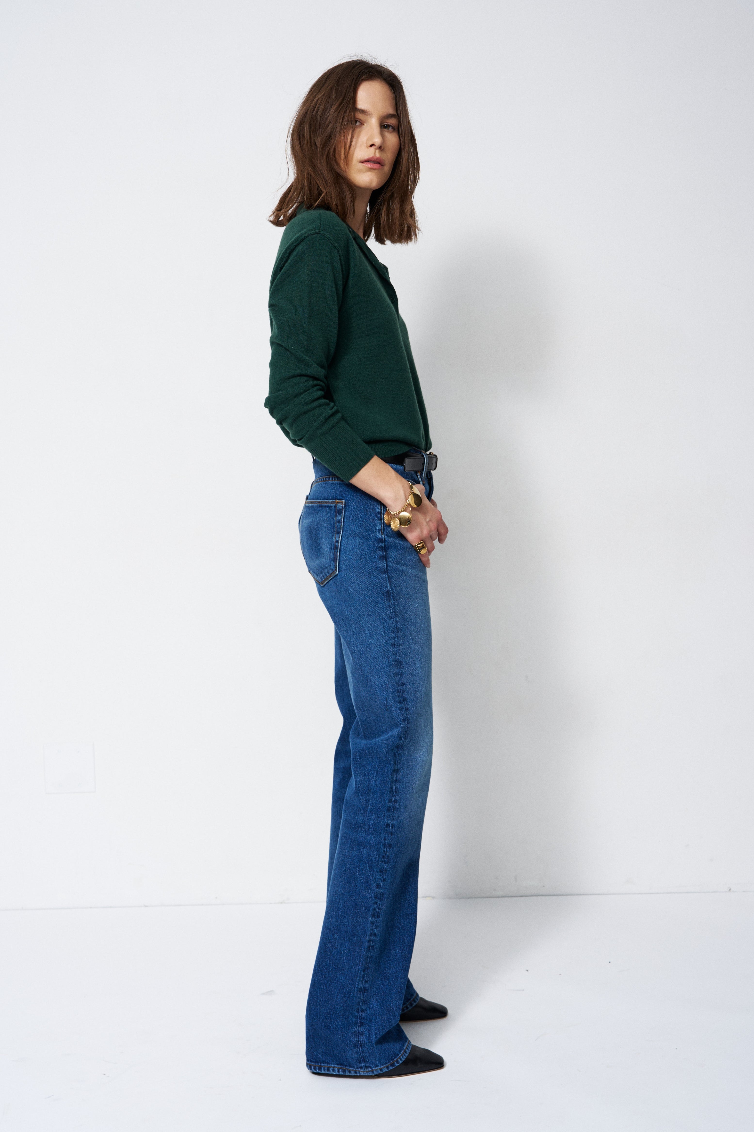 A woman stands in profile wearing a dark green long-sleeve top and blue flare jeans against a plain white studio background.