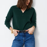 A woman wearing a dark green long-sleeve polo sweater and blue jeans, standing against a plain light gray wall.