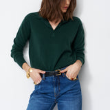 A woman wearing a dark green long-sleeve polo sweater and blue jeans, standing against a plain light gray wall.