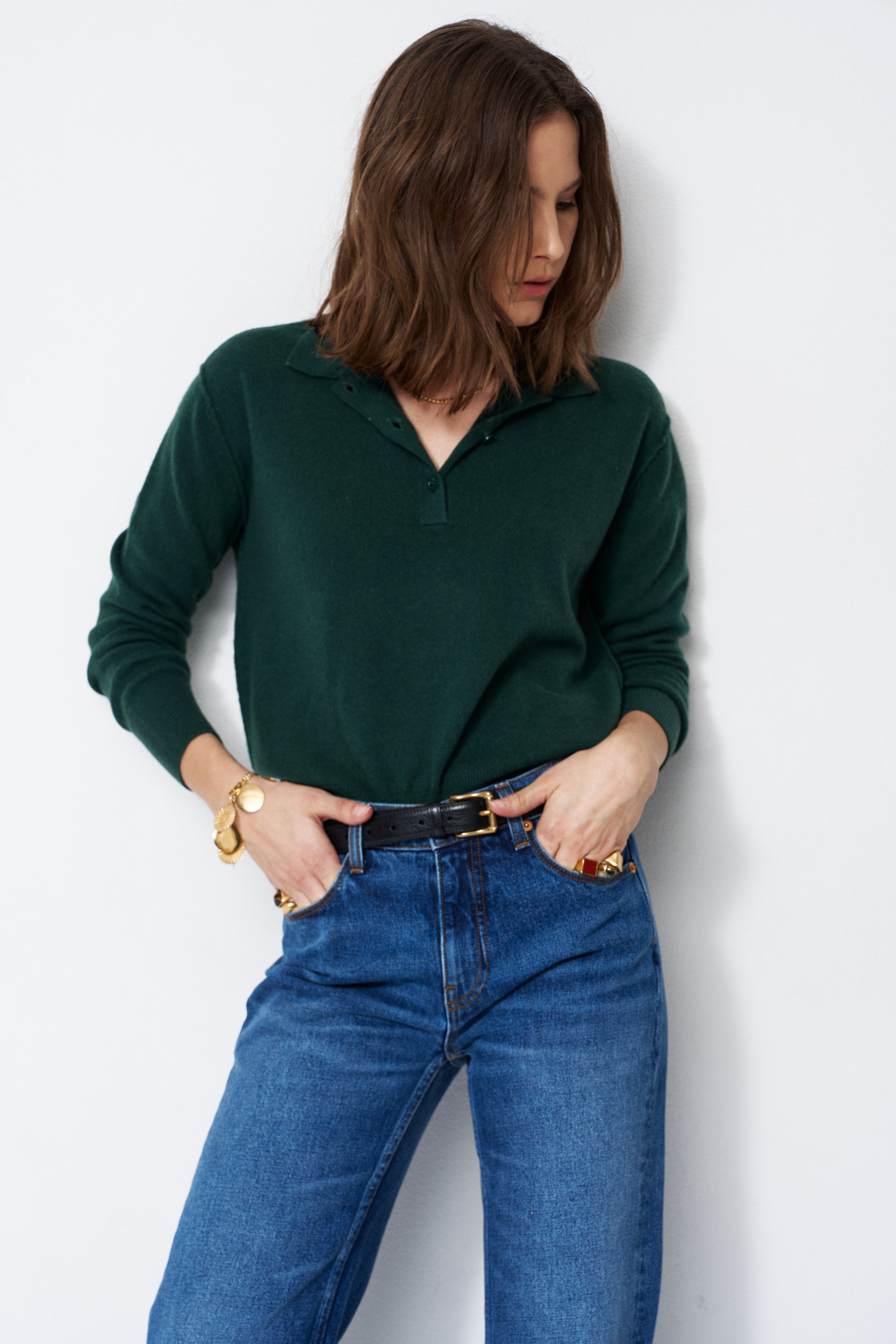 A woman wearing a dark green long-sleeve polo sweater and blue jeans, standing against a plain light gray wall.