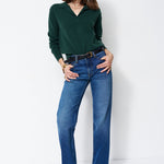 A woman modeling a dark green long-sleeve polo-neck top and blue flare jeans with a black belt, standing against a plain white studio background.