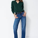 A woman modeling a dark green long-sleeve polo-neck top and blue flare jeans with a black belt, standing against a plain white studio background.