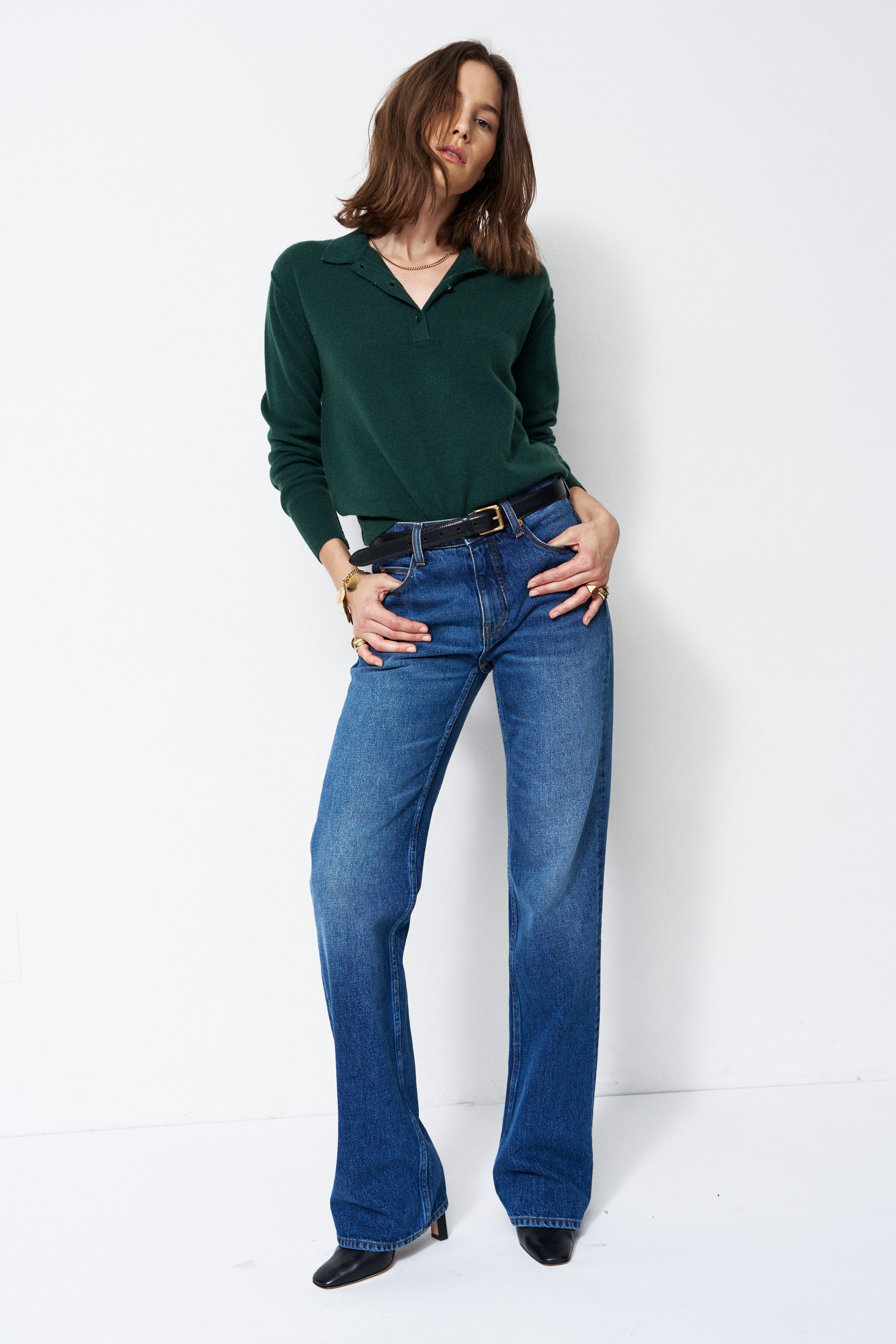 A woman modeling a dark green long-sleeve polo-neck top and blue flare jeans with a black belt, standing against a plain white studio background.