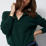 A woman wearing a dark green long-sleeve polo shirt tucked into blue jeans with a black belt, photographed against a plain light gray studio background.