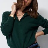 A woman wearing a dark green long-sleeve polo shirt tucked into blue jeans with a black belt, photographed against a plain light gray studio background.