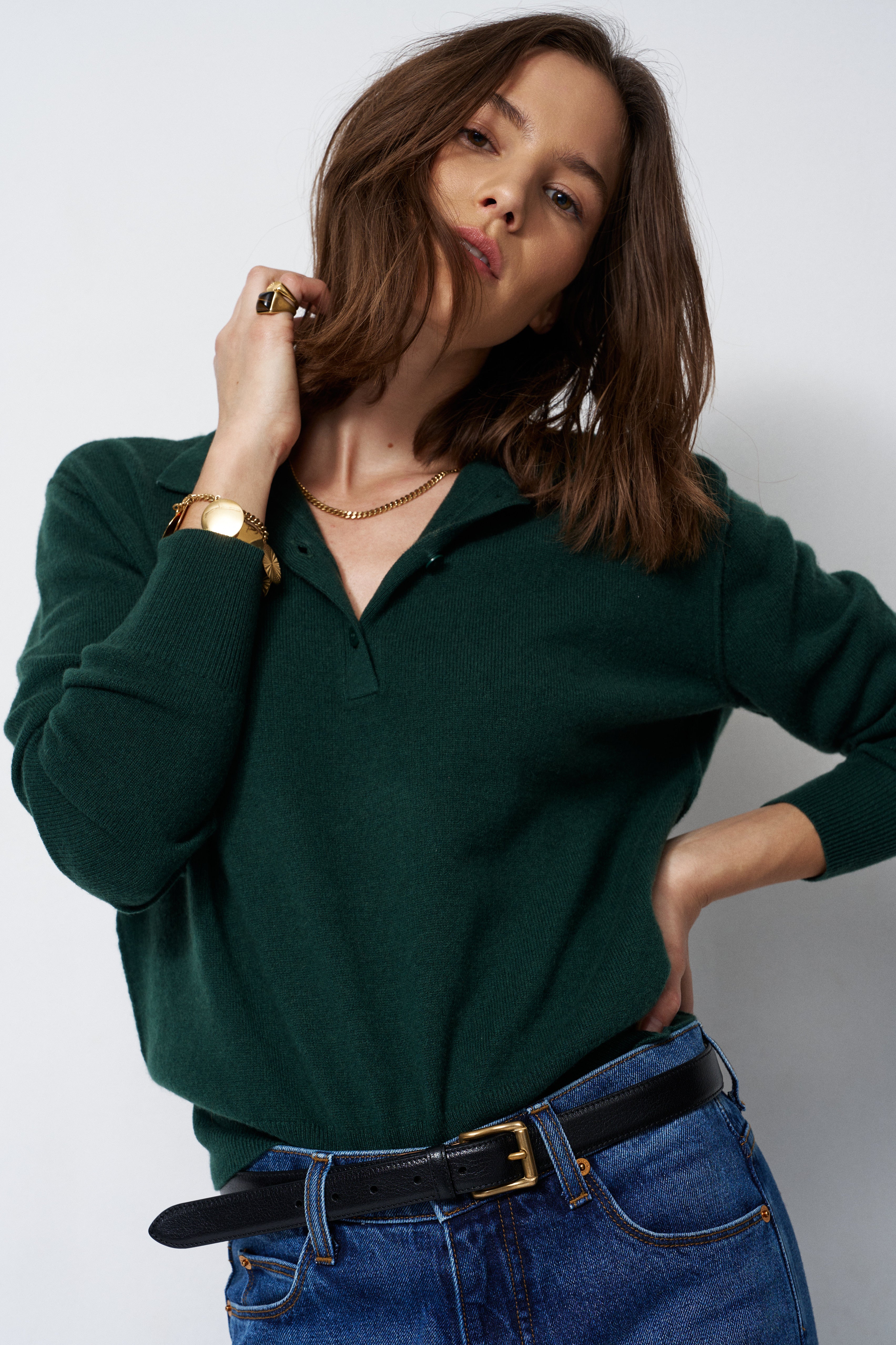 A woman wearing a dark green long-sleeve polo shirt tucked into blue jeans with a black belt, photographed against a plain light gray studio background.