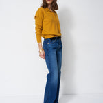 A woman modeling a mustard yellow long-sleeve polo shirt tucked into blue flare jeans, standing against a plain white studio background.