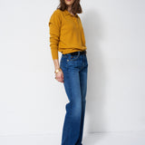 A woman modeling a mustard yellow long-sleeve polo shirt tucked into blue flare jeans, standing against a plain white studio background.