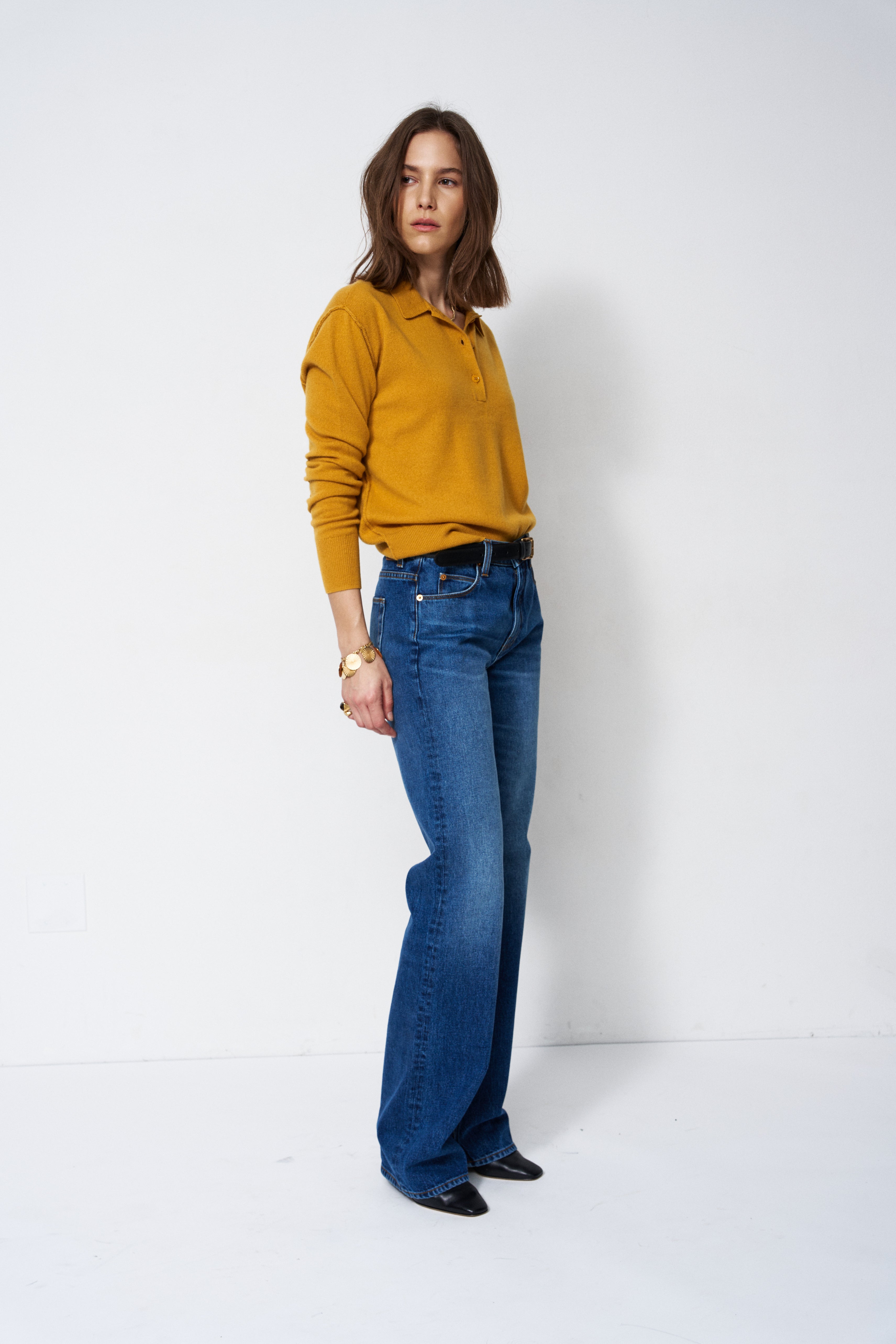 A woman modeling a mustard yellow long-sleeve polo shirt tucked into blue flare jeans, standing against a plain white studio background.