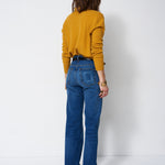 A woman viewed from behind wearing a mustard yellow long-sleeve top and blue high-waisted flare jeans, standing against a plain white studio background.