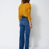 A woman viewed from behind wearing a mustard yellow long-sleeve top and blue high-waisted flare jeans, standing against a plain white studio background.