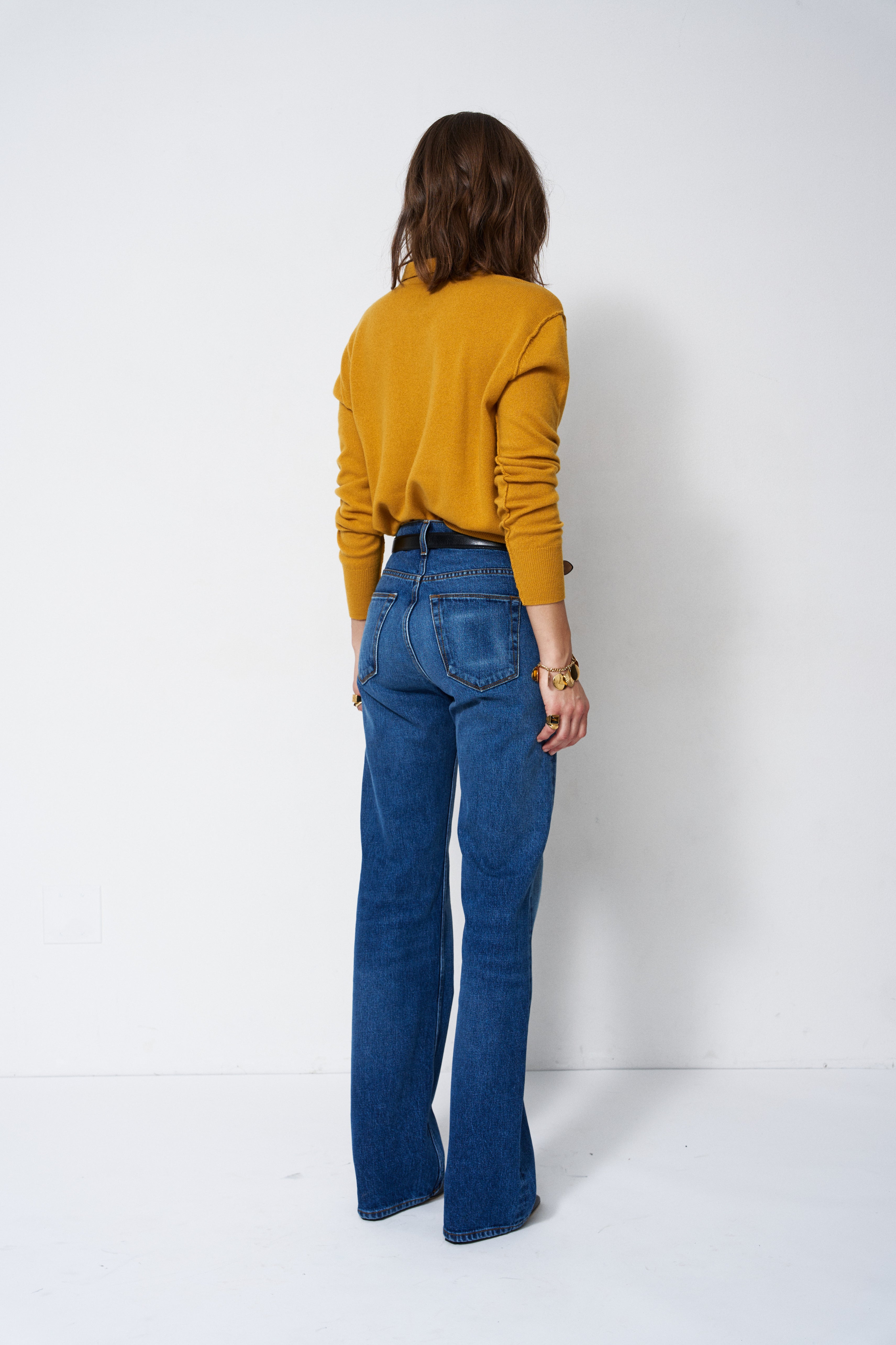 A woman viewed from behind wearing a mustard yellow long-sleeve top and blue high-waisted flare jeans, standing against a plain white studio background.