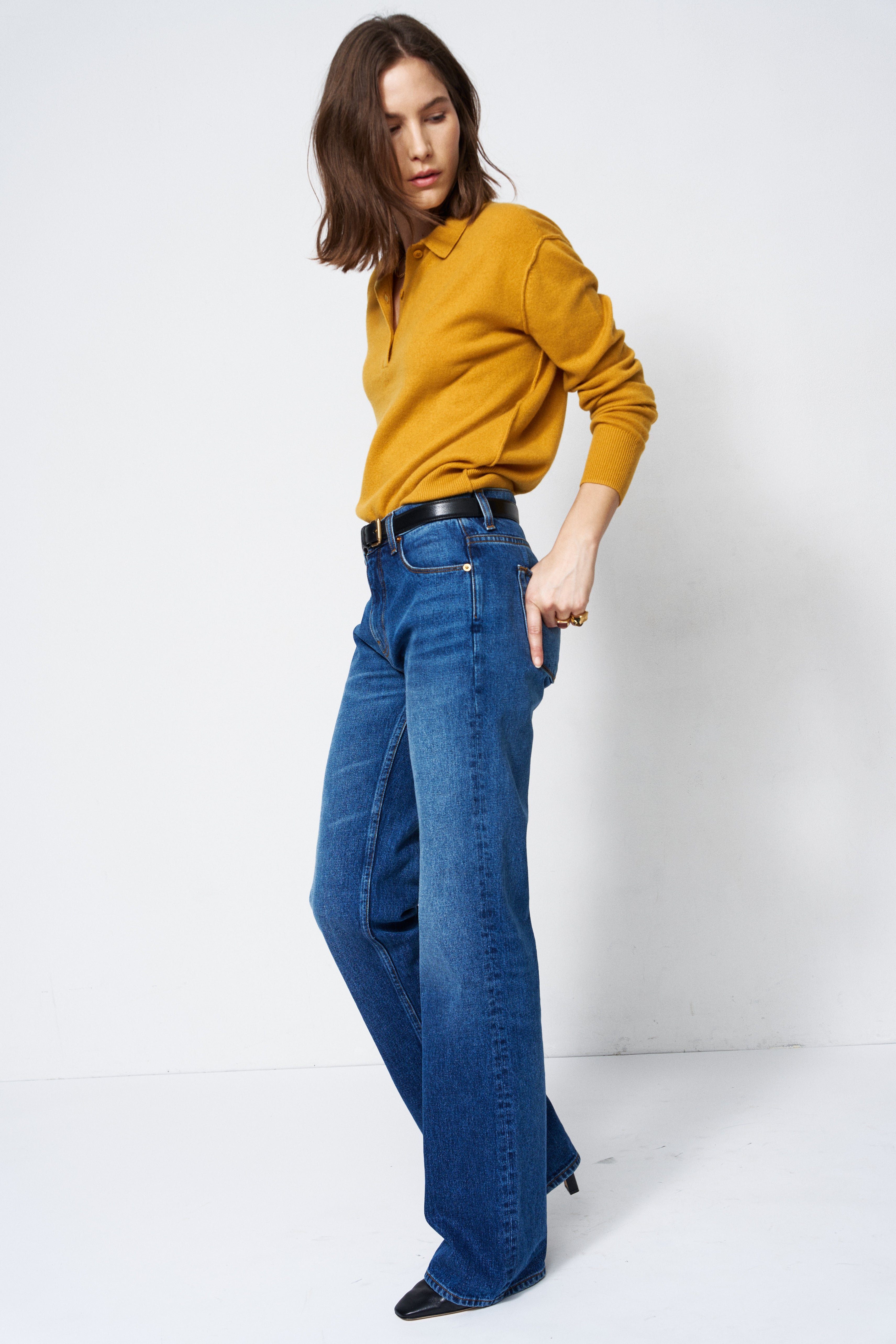 A woman modeling a mustard yellow long-sleeve knit top and high-waisted, wide-leg blue jeans with a black belt, standing against a plain white studio background.
