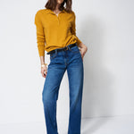 A woman modeling a mustard yellow long-sleeve polo shirt tucked into high-waisted, wide-leg blue jeans with a black belt, standing against a plain white studio background.