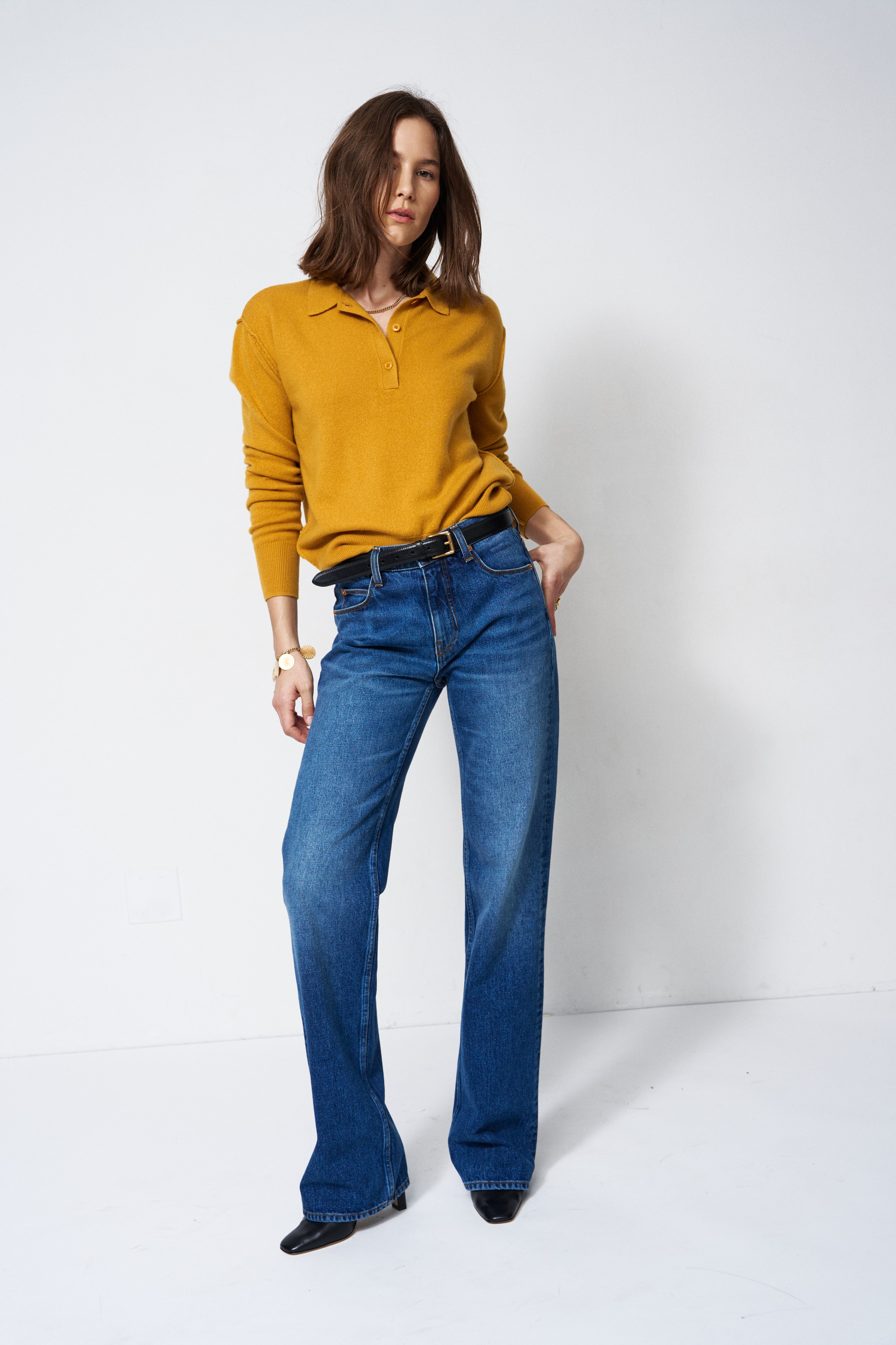A woman modeling a mustard yellow long-sleeve polo shirt tucked into high-waisted, wide-leg blue jeans with a black belt, standing against a plain white studio background.