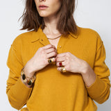 Woman wearing a mustard yellow sweater with jewelry on a plain background