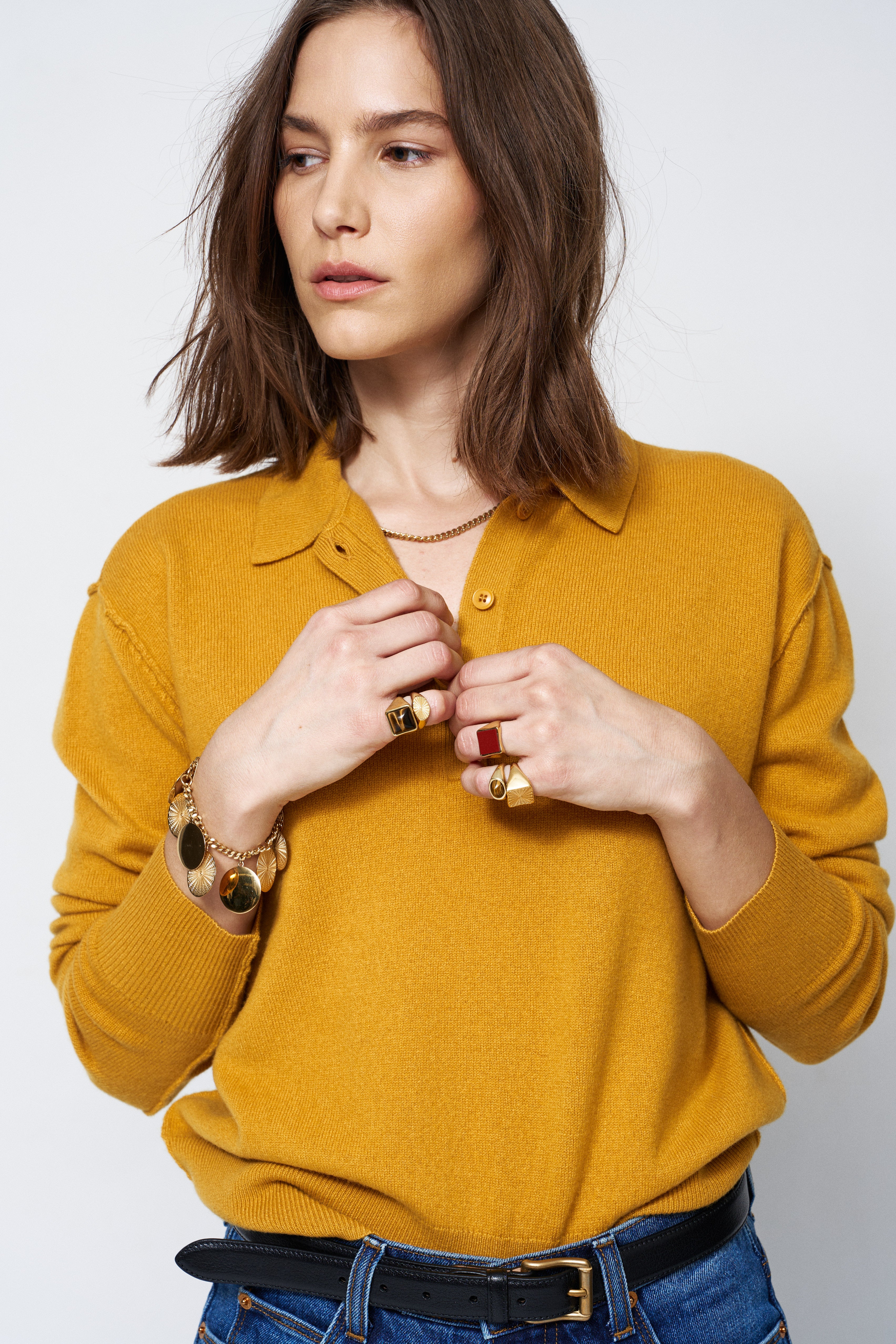 Woman wearing a mustard yellow sweater with jewelry on a plain background