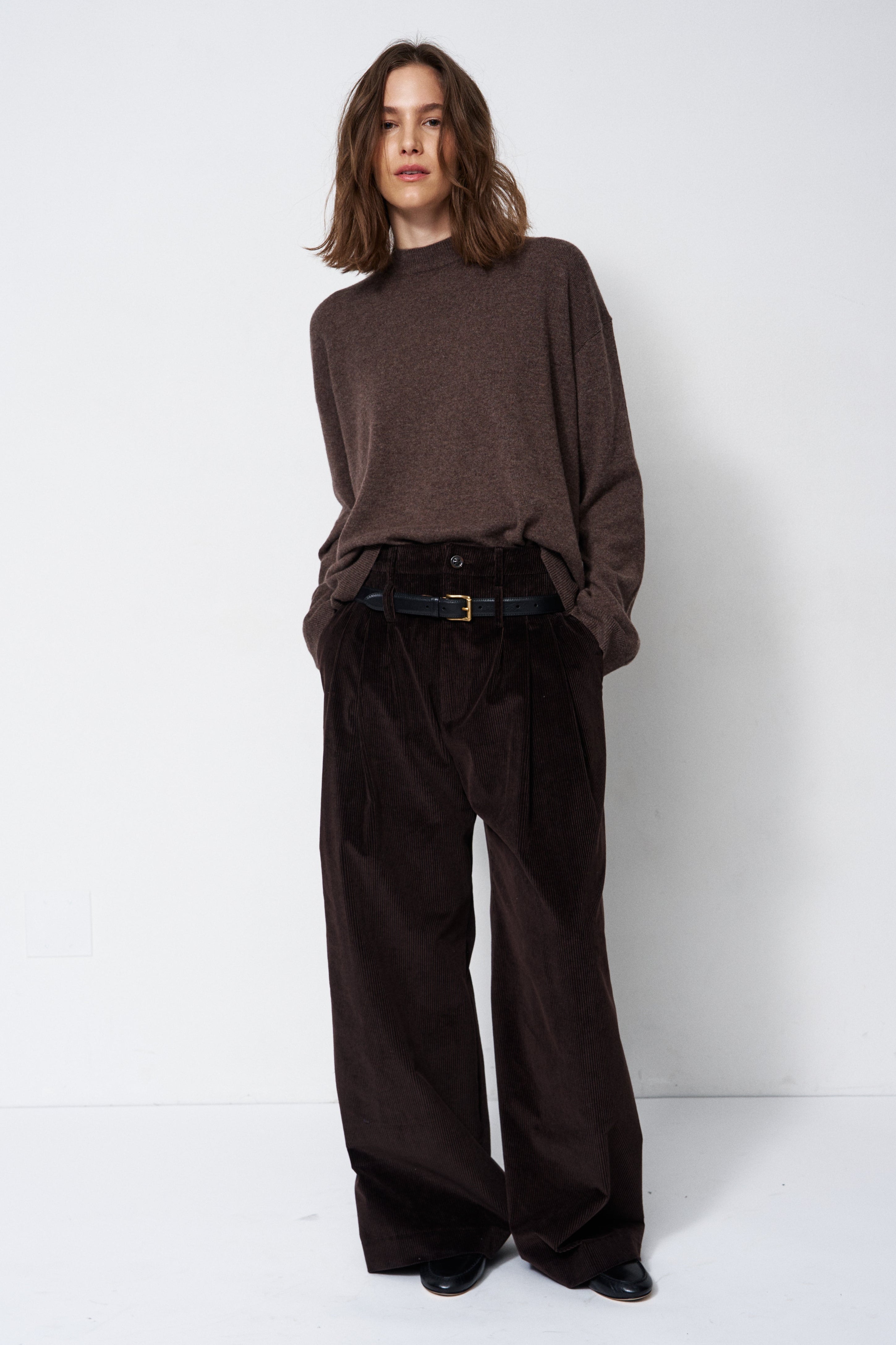 A model stands against a plain white studio background wearing a dark brown mock-neck knit sweater, dark brown wide-leg corduroy trousers, and a black belt.