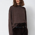 A woman wearing a dark brown mock-neck knit sweater and dark brown corduroy trousers, styled with a black belt, standing against a light gray studio background.