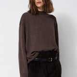 A woman wearing a dark brown mock-neck knit sweater and dark brown corduroy trousers, styled with a black belt, standing against a light gray studio background.