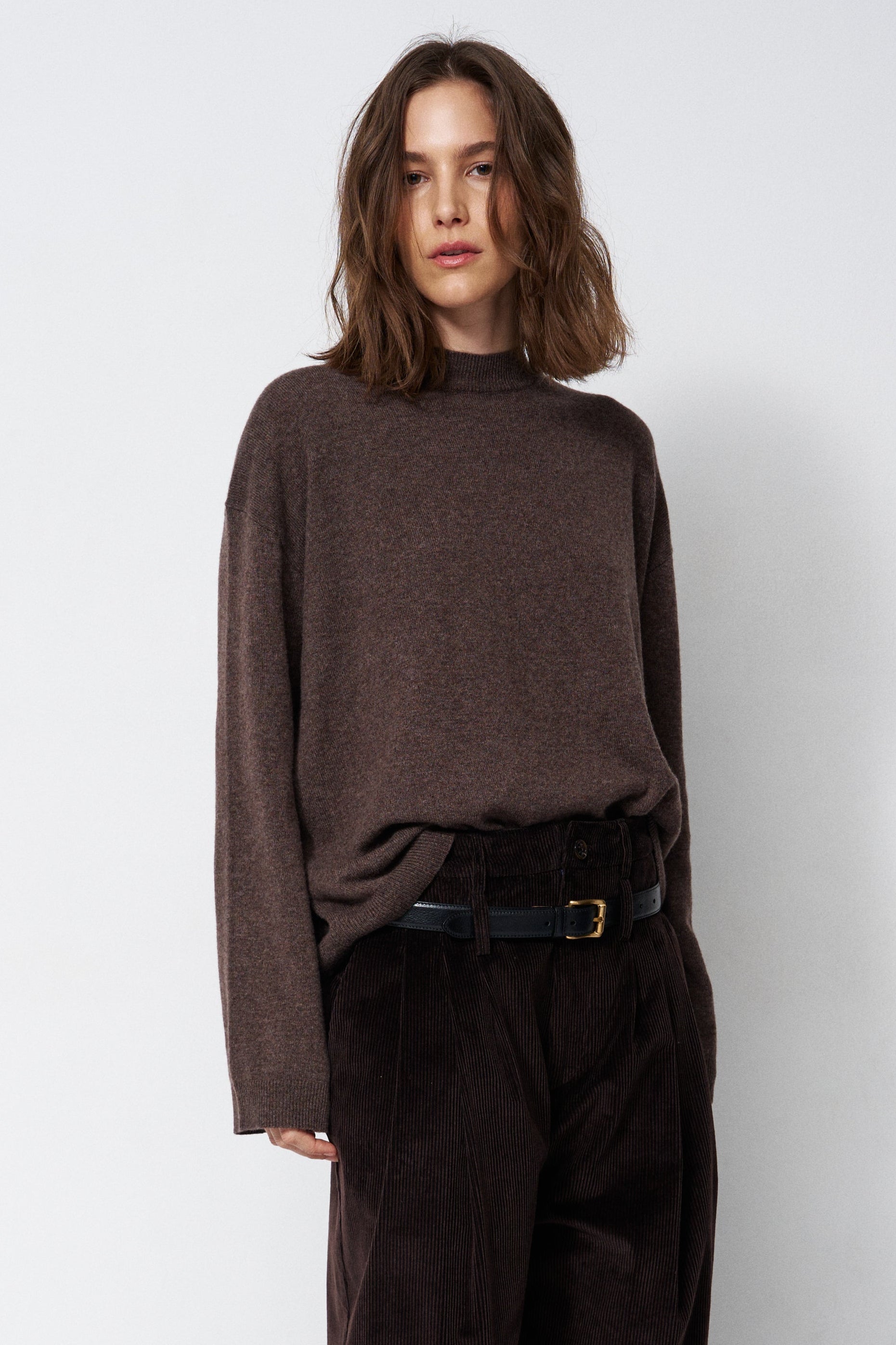 A woman wearing a dark brown mock-neck knit sweater and dark brown corduroy trousers, styled with a black belt, standing against a light gray studio background.