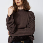 A woman wearing a dark brown knit sweater and dark brown corduroy trousers with a black belt, posing against a light gray studio background.