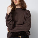A woman wearing a dark brown knit sweater and dark brown corduroy trousers with a black belt, posing against a light gray studio background.