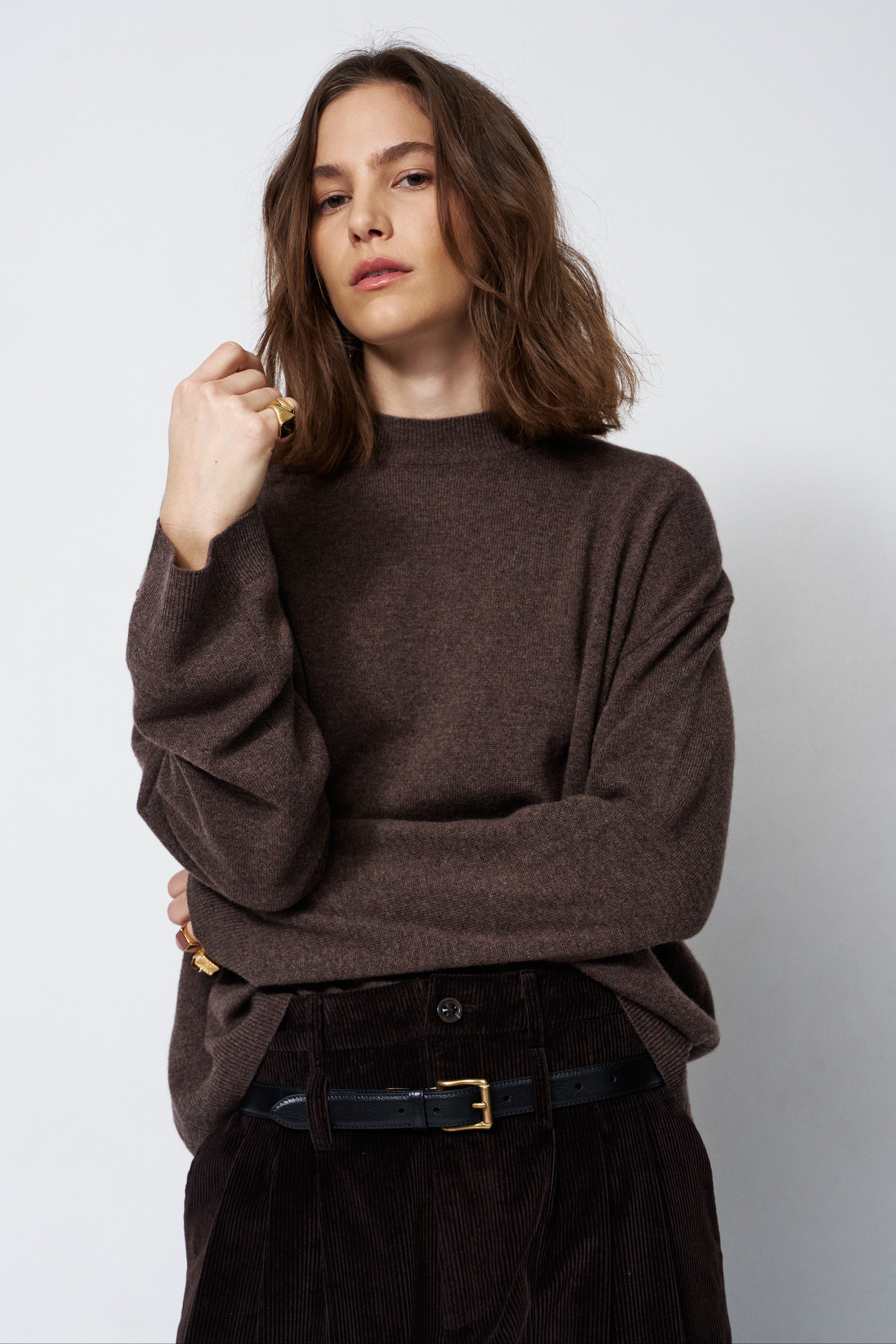 A woman wearing a dark brown knit sweater and dark brown corduroy trousers with a black belt, posing against a light gray studio background.