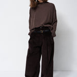 A woman modeling a dark brown knit mock-neck sweater and matching dark brown wide-leg corduroy trousers, styled with a black belt and black shoes, against a plain white studio background.
