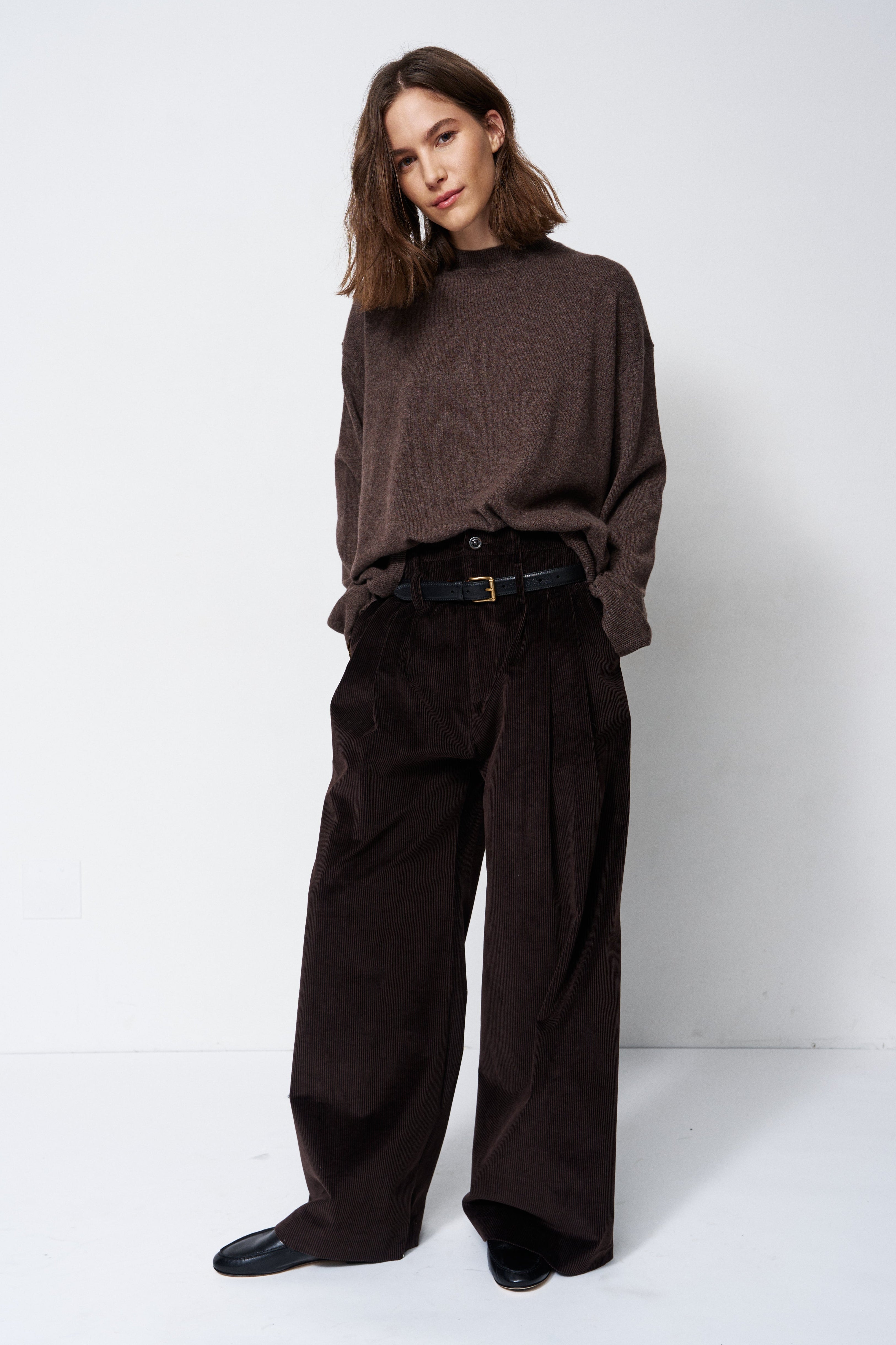 A woman modeling a dark brown knit mock-neck sweater and matching dark brown wide-leg corduroy trousers, styled with a black belt and black shoes, against a plain white studio background.