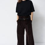 A woman stands against a plain white studio background wearing a loose black short-sleeve T-shirt and very wide-leg dark brown corduroy trousers with a black belt.