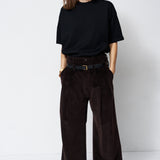 A woman stands against a plain white studio background wearing a loose black short-sleeve T-shirt and very wide-leg dark brown corduroy trousers with a black belt.