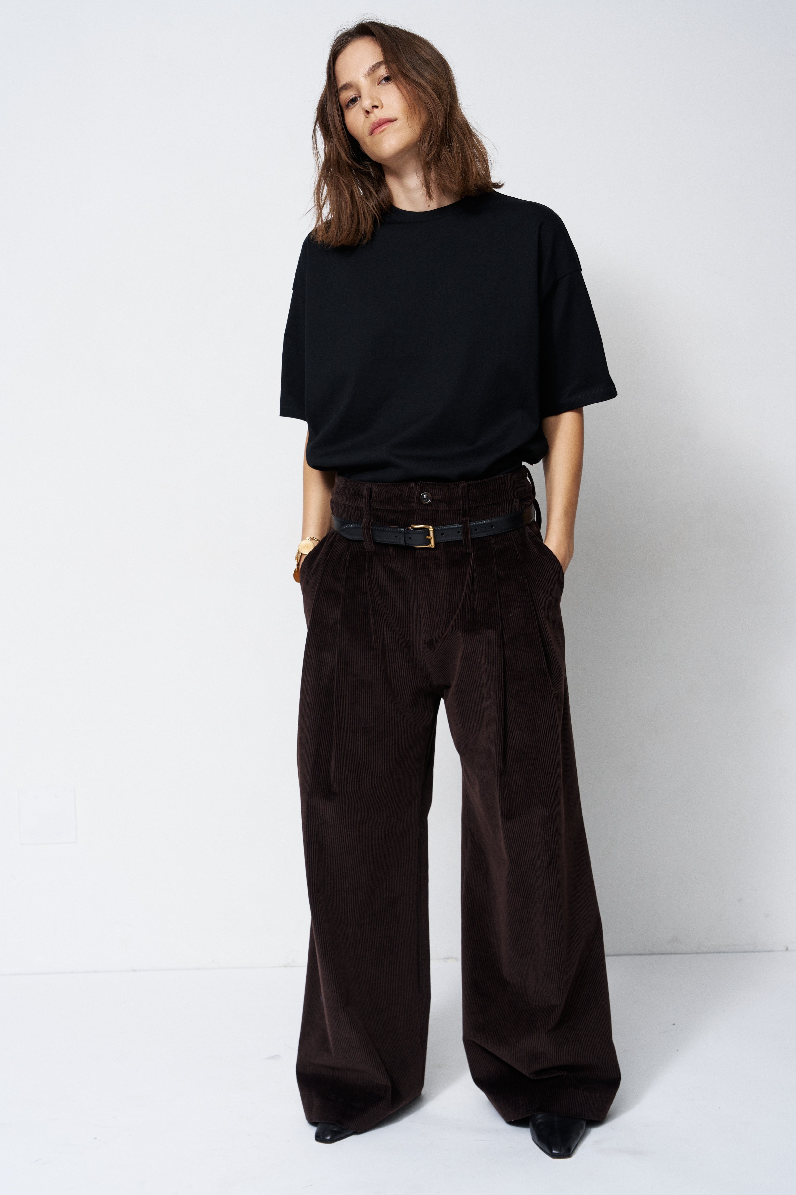 A woman stands against a plain white studio background wearing a loose black short-sleeve T-shirt and very wide-leg dark brown corduroy trousers with a black belt.