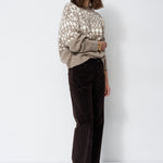 A woman stands against a plain white studio background wearing a beige and white patterned knit sweater, dark brown wide-leg corduroy pants, and black pointed shoes.