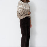 A woman stands against a plain white studio background wearing a beige and white patterned knit sweater, dark brown wide-leg corduroy pants, and black pointed shoes.
