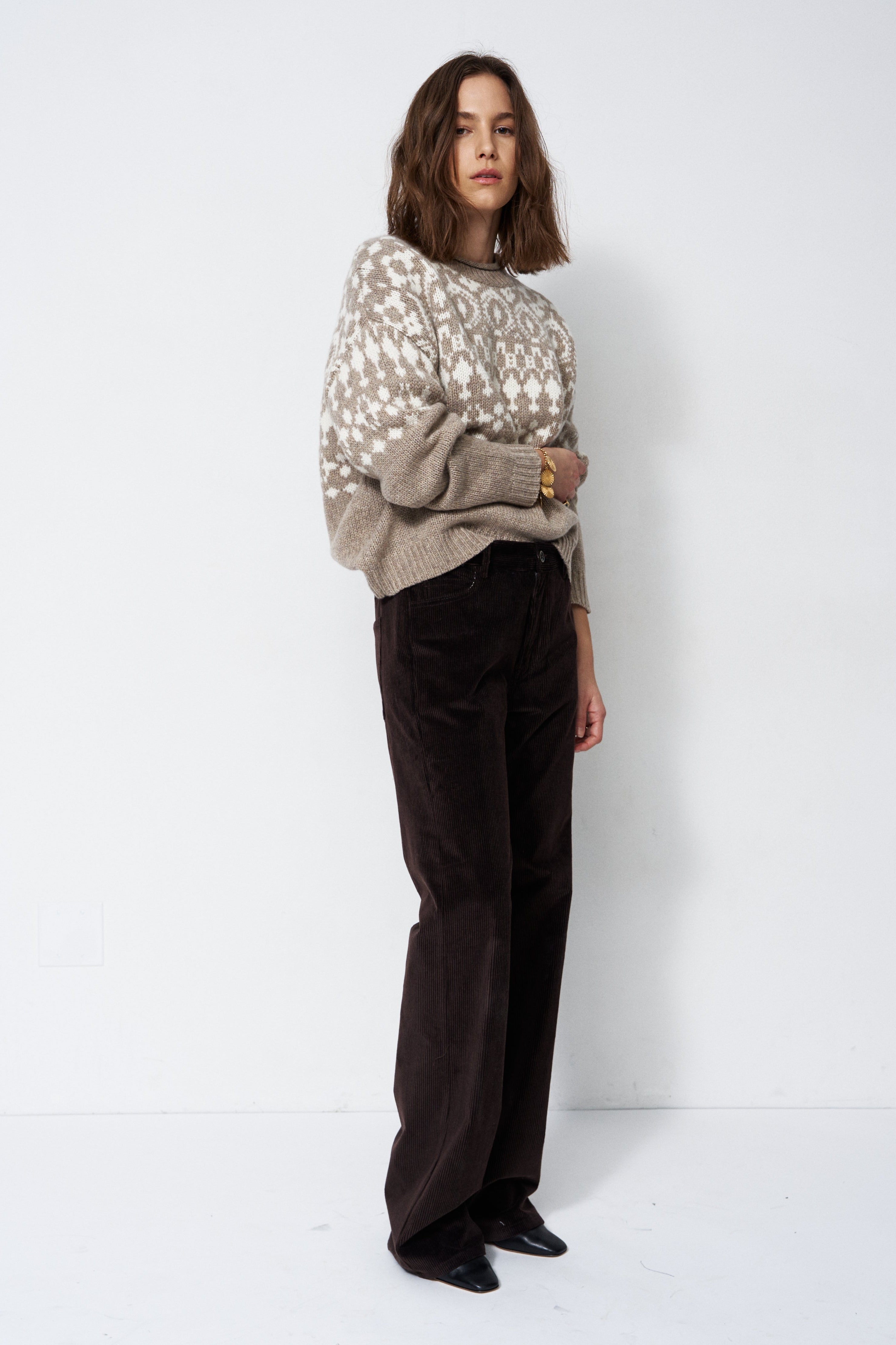 A woman stands against a plain white studio background wearing a beige and white patterned knit sweater, dark brown wide-leg corduroy pants, and black pointed shoes.
