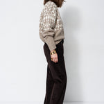 A woman stands in profile wearing a beige knit sweater with a white geometric pattern and dark brown wide-leg trousers, photographed against a plain white studio background.