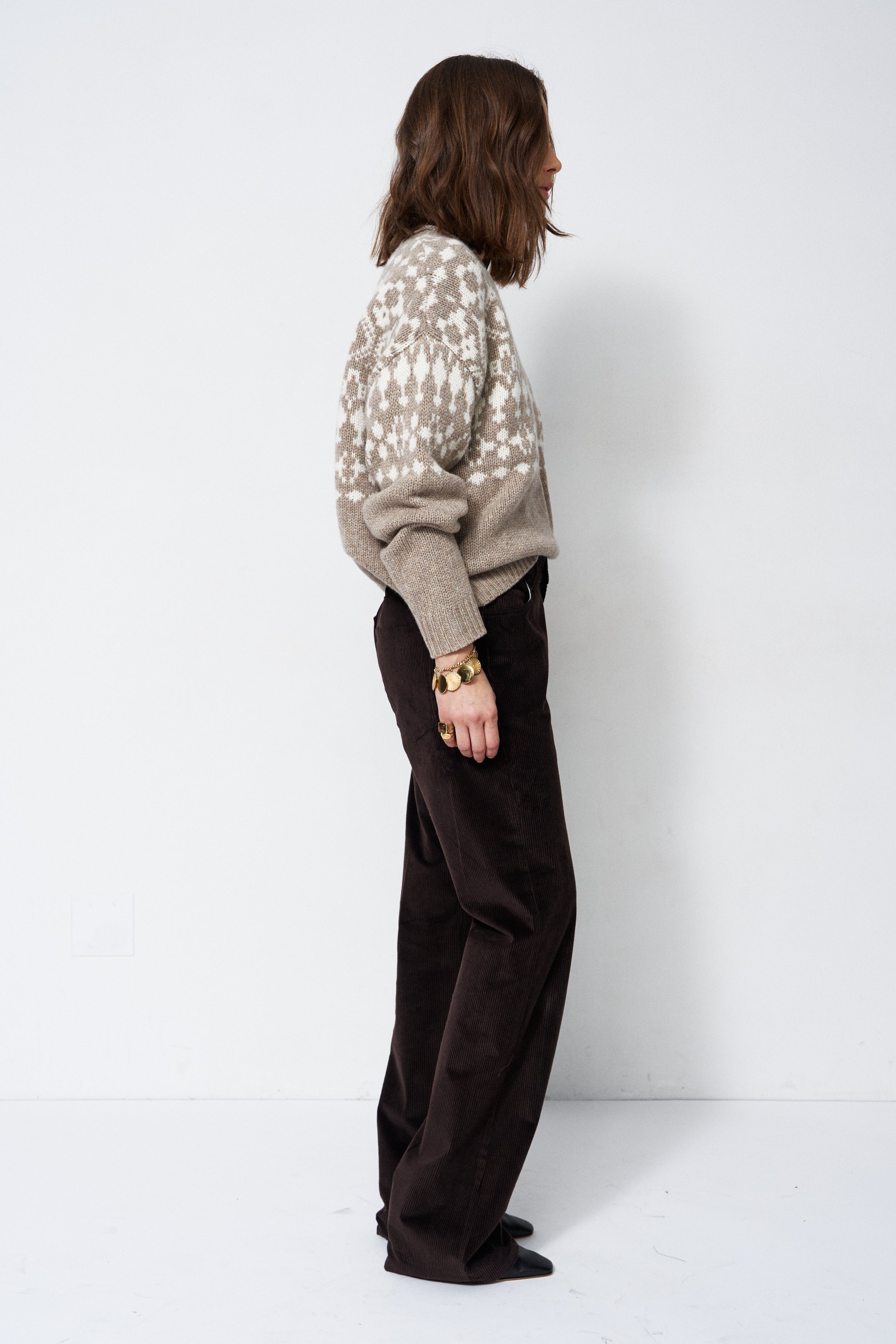 A woman stands in profile wearing a beige knit sweater with a white geometric pattern and dark brown wide-leg trousers, photographed against a plain white studio background.
