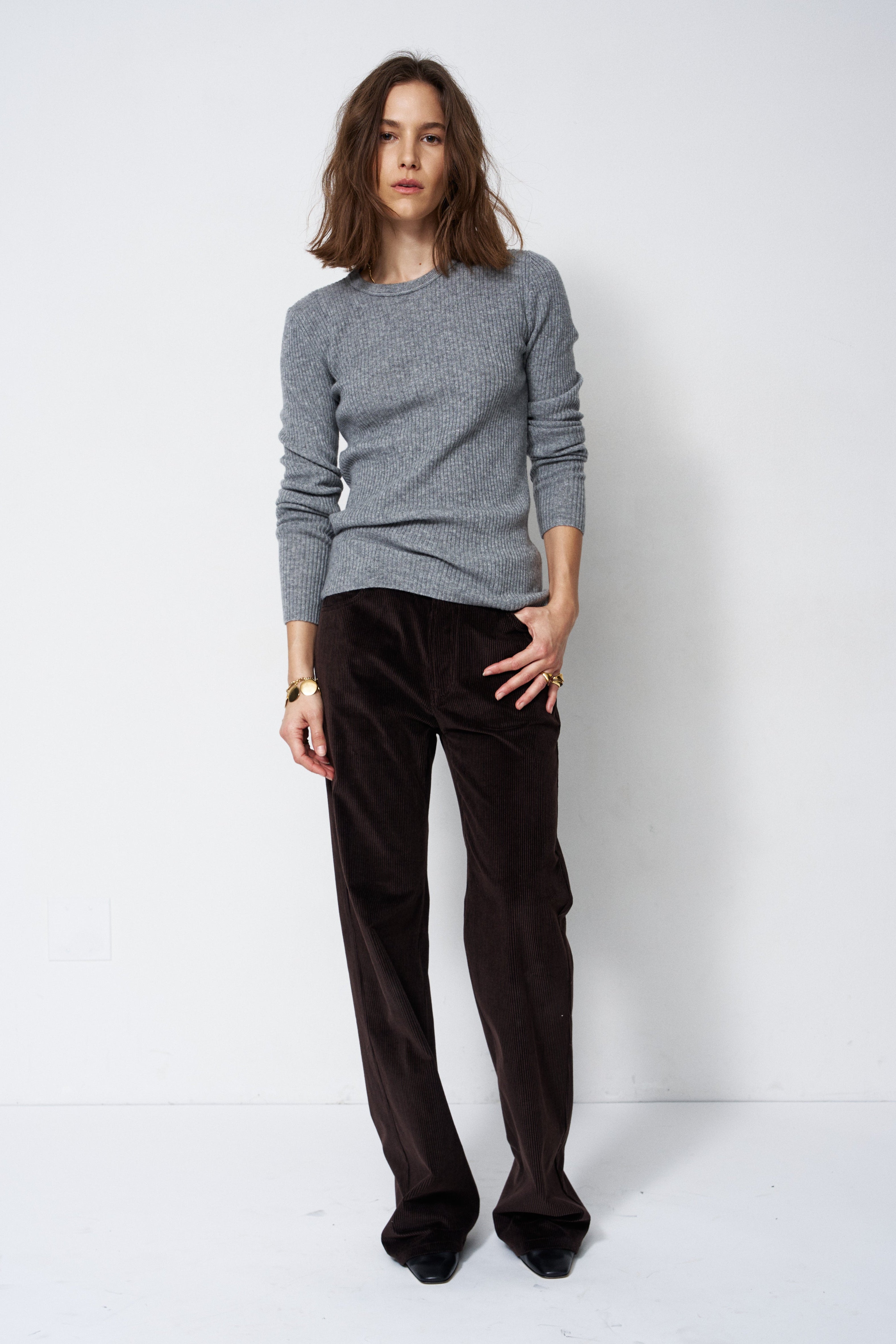 A woman modeling a light gray knit crewneck sweater and dark brown wide-leg corduroy trousers against a plain white studio background.