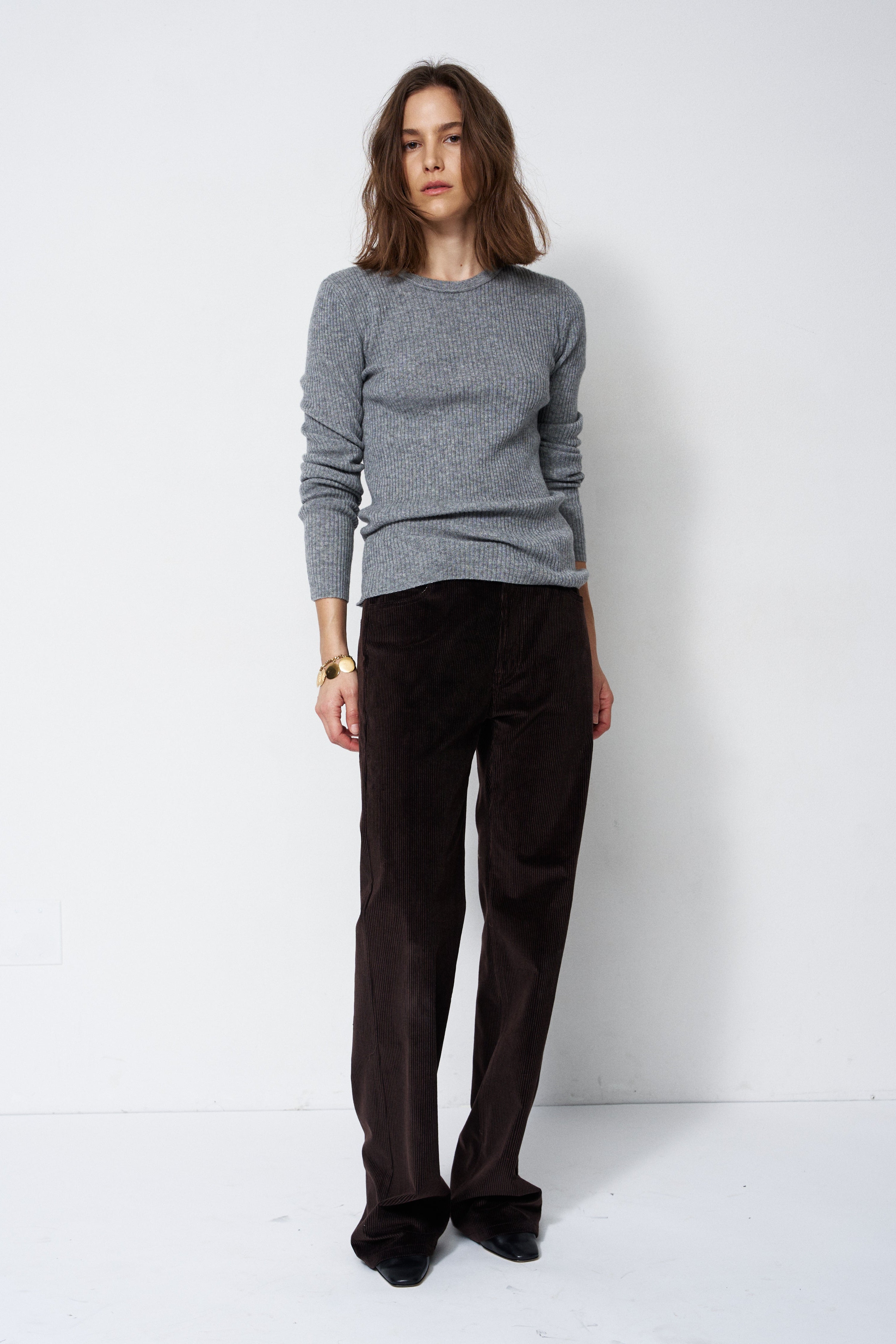A woman modeling a gray knit crewneck sweater and dark brown corduroy trousers against a plain white studio background.