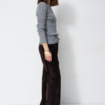 A woman stands in profile wearing a light gray long-sleeve knit sweater and dark brown wide-leg trousers against a plain white studio background.