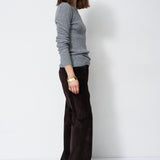 A woman stands in profile wearing a light gray long-sleeve knit sweater and dark brown wide-leg trousers against a plain white studio background.